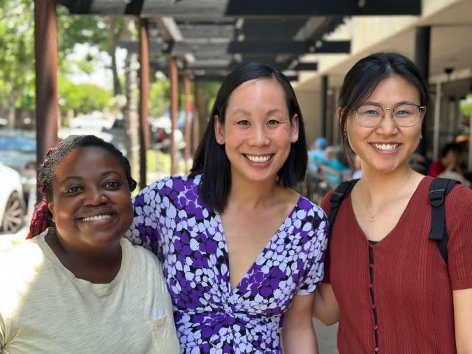 Three smiling women standing close on a sunny street market; middle in purple floral dress.