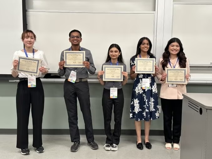 Five smiling young adults in a classroom holding certificates in front of a whiteboard